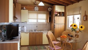 A compact kitchen with wooden cabinets, a built-in oven, a sink, and a dining area with wicker chairs and a glass table. - Amalia House Garden (Pefki)