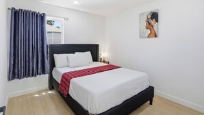 A hotel room with a bed, a window with curtains, a wall-mounted painting, and a bedside table with a lamp.