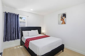A hotel room with a bed, a window with curtains, a wall-mounted painting, and a bedside table with a lamp.