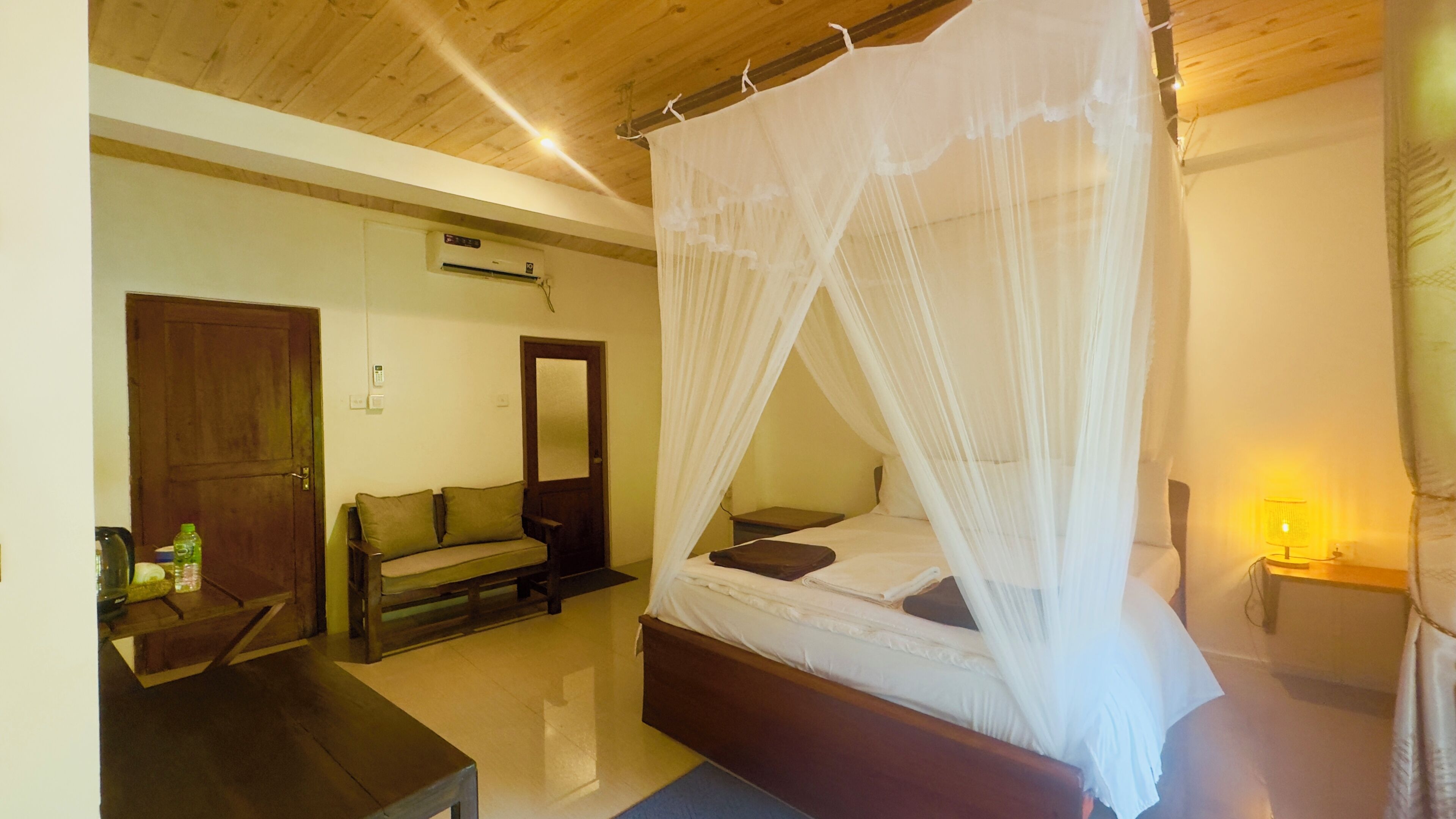 A bedroom with a canopy bed, a wooden ceiling, a sofa, a bedside table, and a lamp.