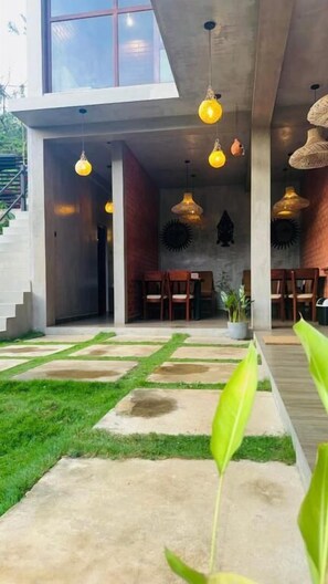 A modern outdoor seating area with concrete flooring, green grass, and hanging light fixtures. - Wild Resort Ella (Ella)