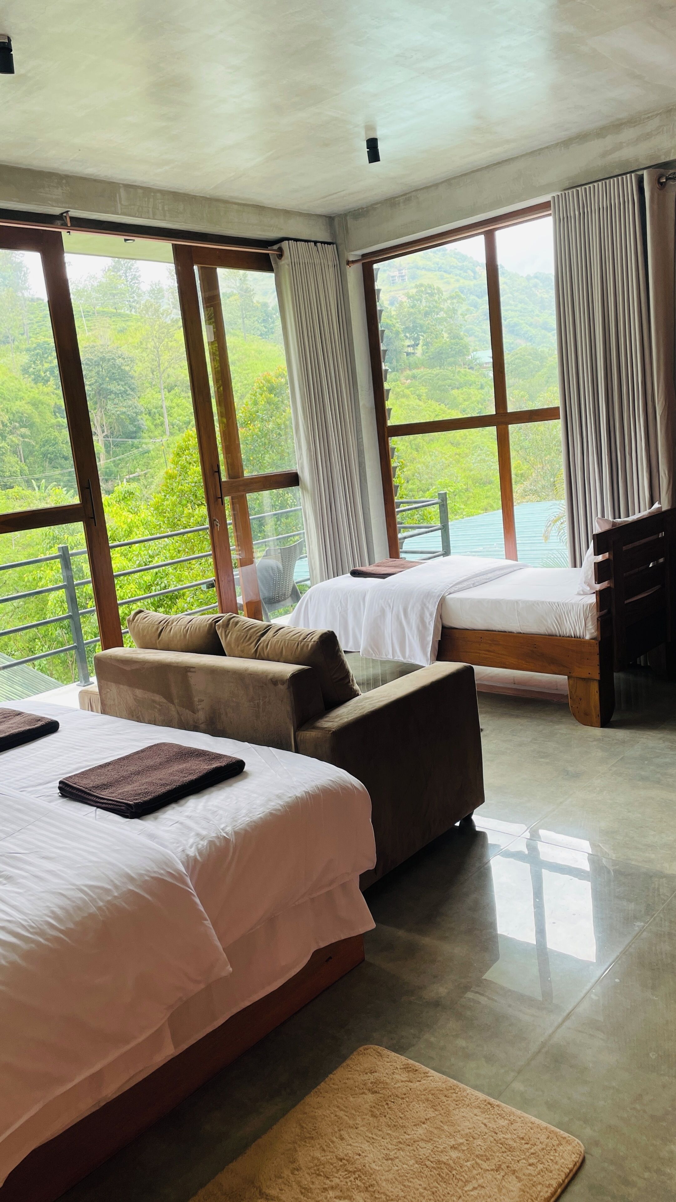 A hotel room with two beds, a sofa, and large windows offering a view of greenery.