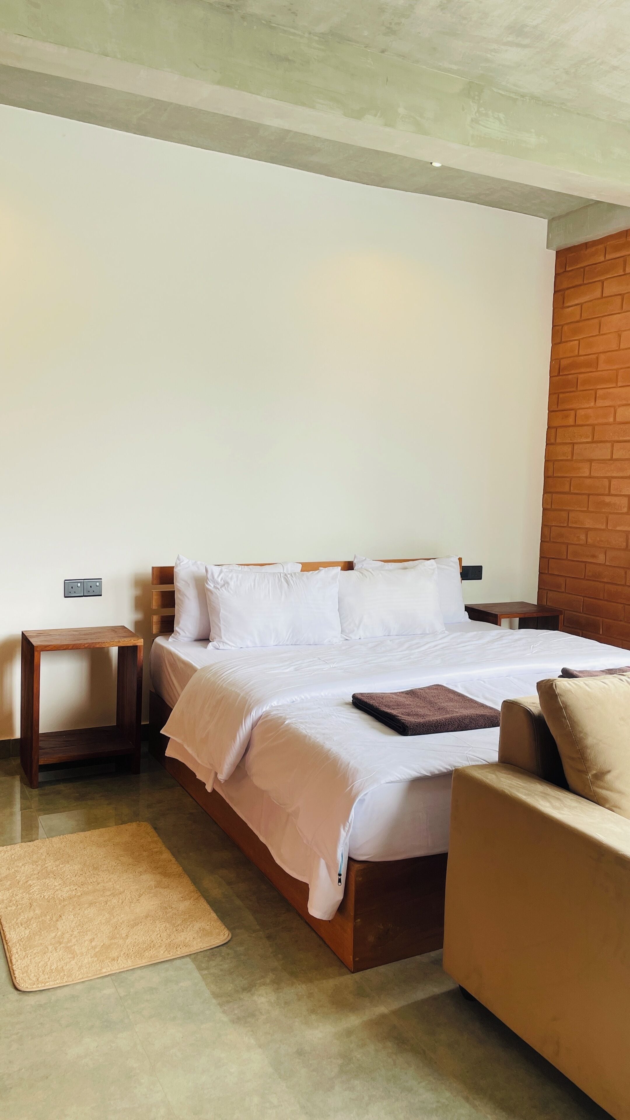 A hotel room with a bed, a wooden nightstand, a small table, a brown leather sofa, and a brick accent wall.