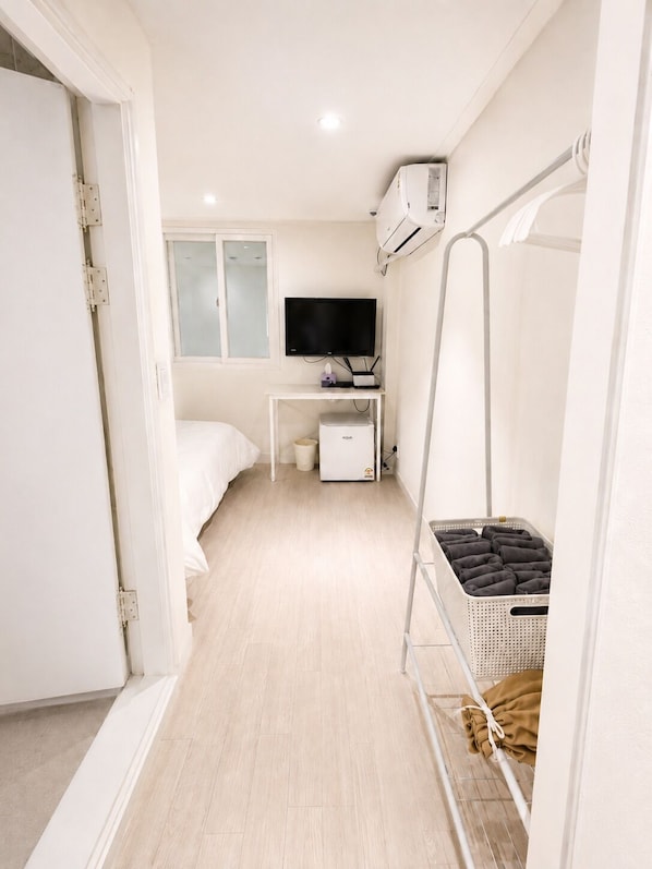 Une chambre d’hôtel de taille réduite, comprenant un lit, un bureau avec une télévision, un appareil de climatisation et un panier à linge.