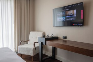 Habitación de hotel con televisión montada en la pared mostrando información del hotel, un escritorio de madera, una silla acolchada y una ventana con cortinas.