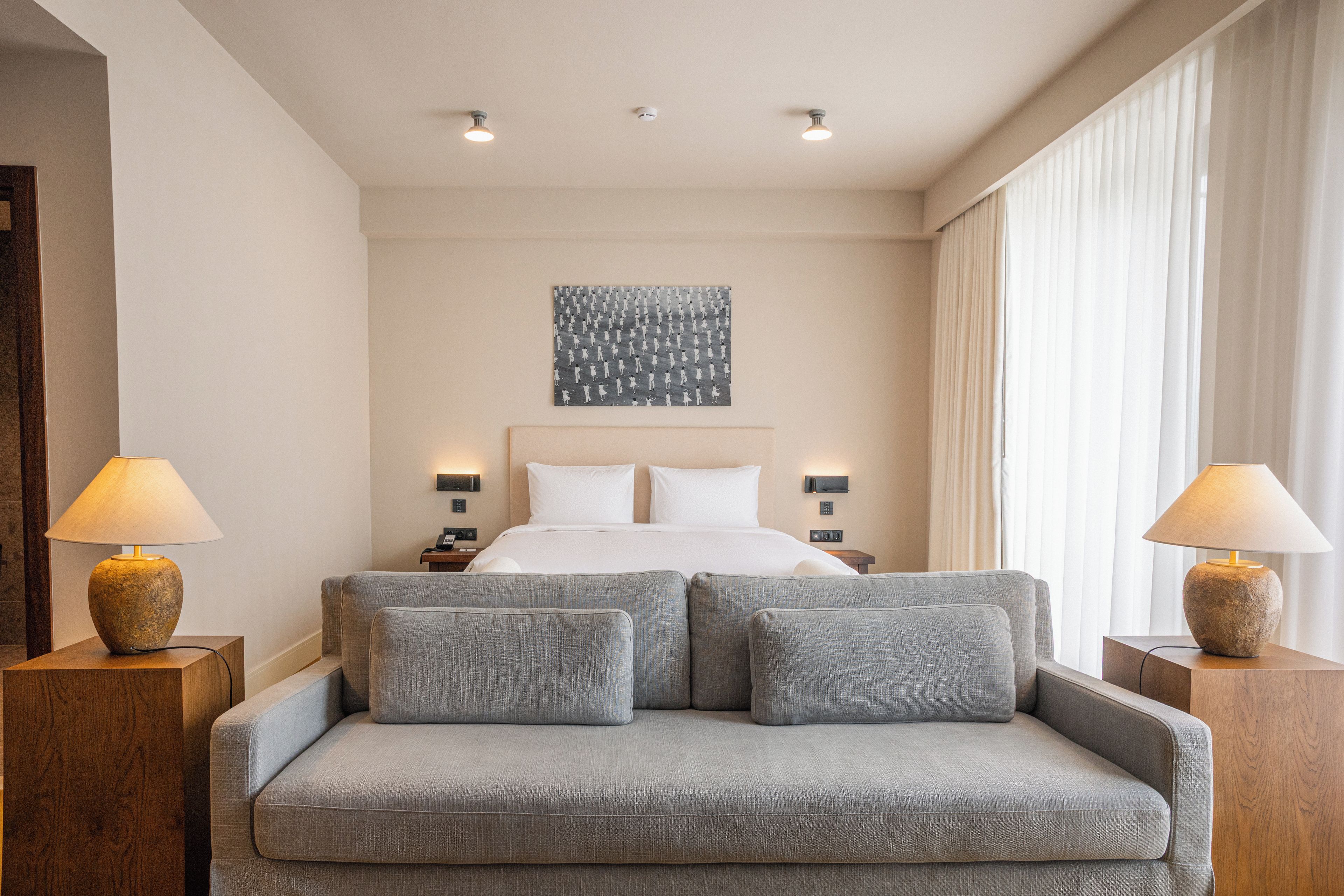 Quarto de hotel com uma cama, um sofá cinzento, duas mesas de cabeceira e uma obra de arte na parede.