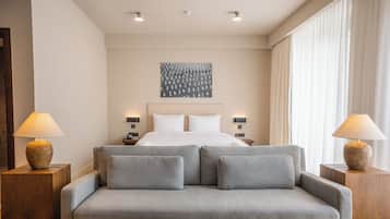 Quarto de hotel com uma cama, um sofá cinzento, duas mesas de cabeceira e uma obra de arte na parede.