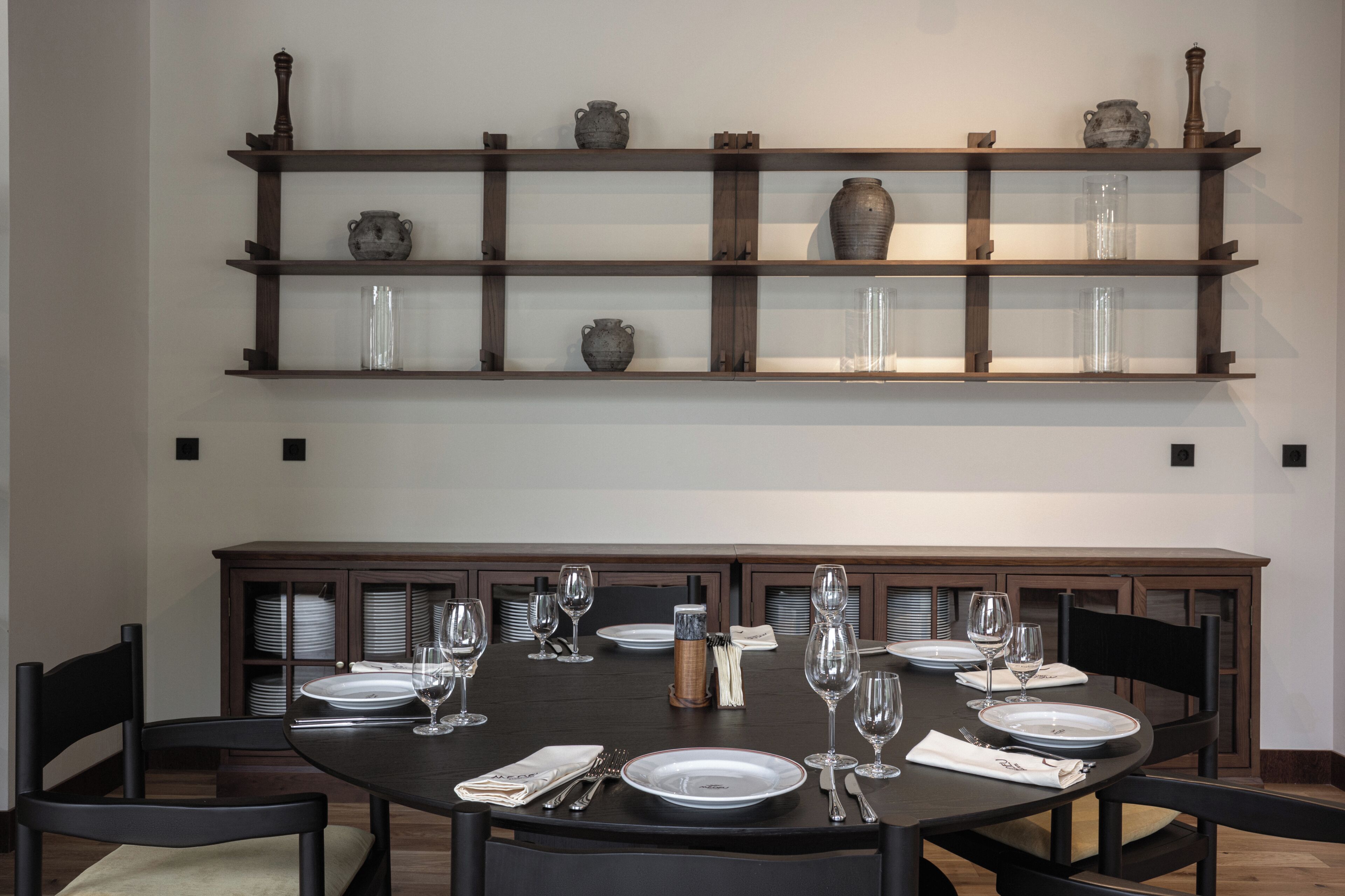 A dining area with a round table set for six, surrounded by chairs. Above the table, there is a wooden shelf with decorative vases and glassware.
