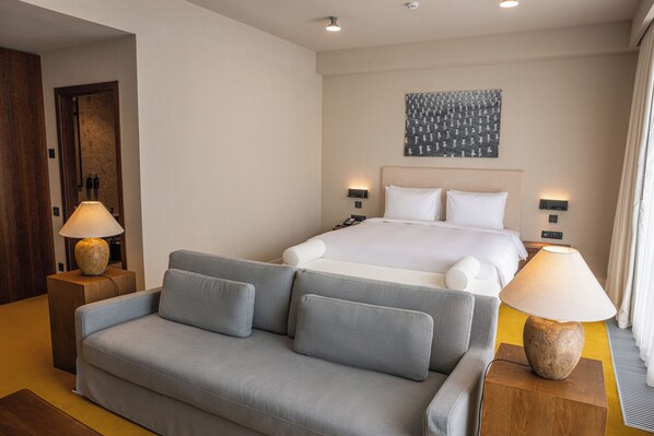 Habitación de hotel con cama, sofá gris, dos lámparas de noche, una obra de arte en la pared y una puerta de madera.