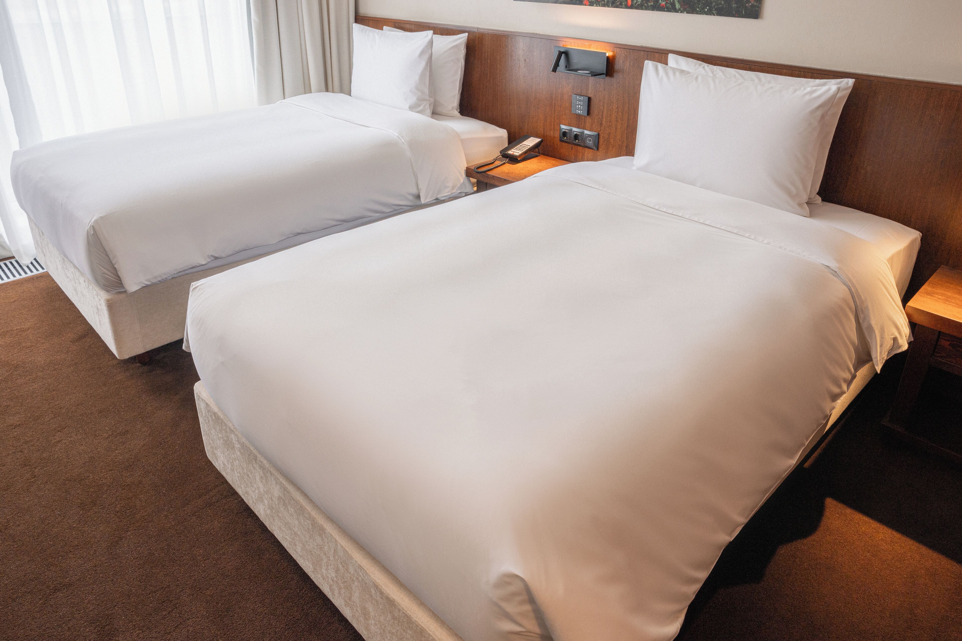 Ein Hotelzimmer mit zwei Betten, einem hölzernen Kopfteil, einem Nachttisch mit Lampe und einem an der Wand befestigten Telefon.