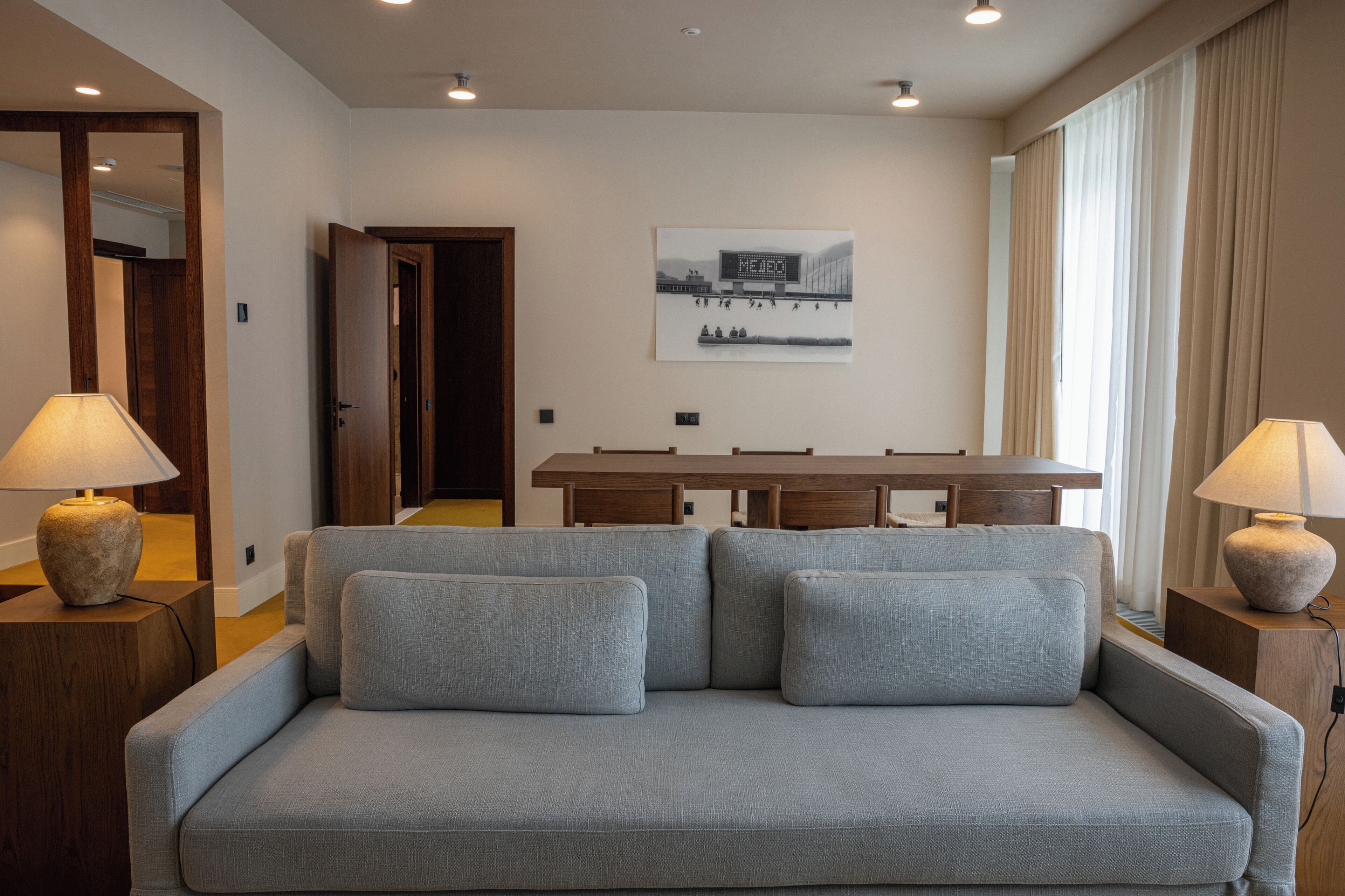 Una habitación de hotel moderna con un sofá gris, mesitas de madera, un escritorio y un ventanal con cortinas.