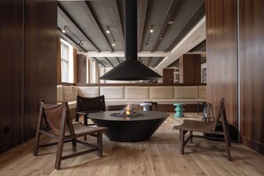 A modern interior with a central fire pit, wooden furniture, and a high ceiling with exposed beams.