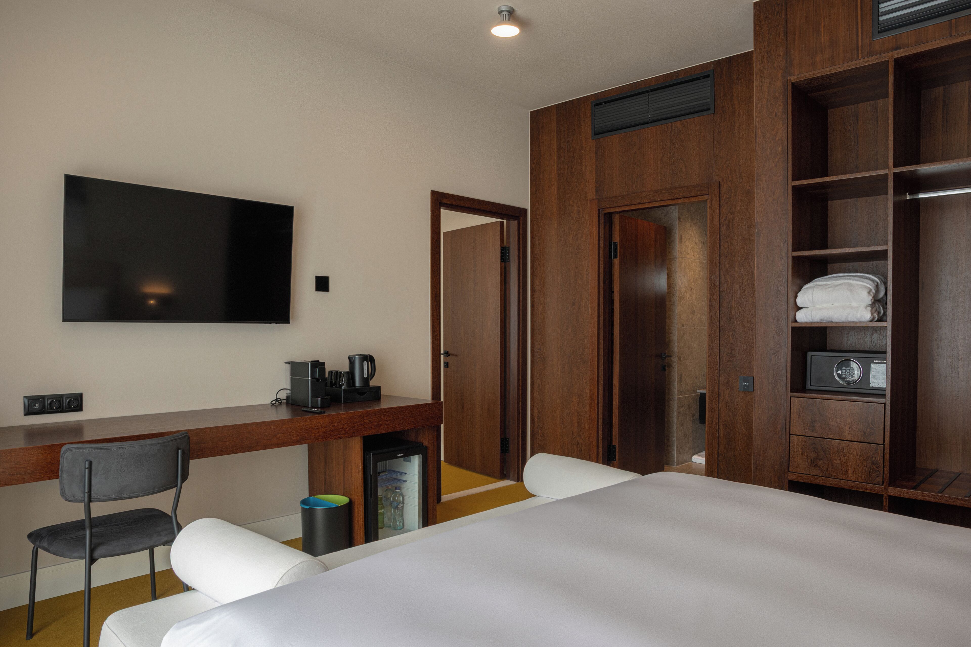 A hotel room with a bed, a desk with a chair, a television, a wardrobe, and a small kitchenette.