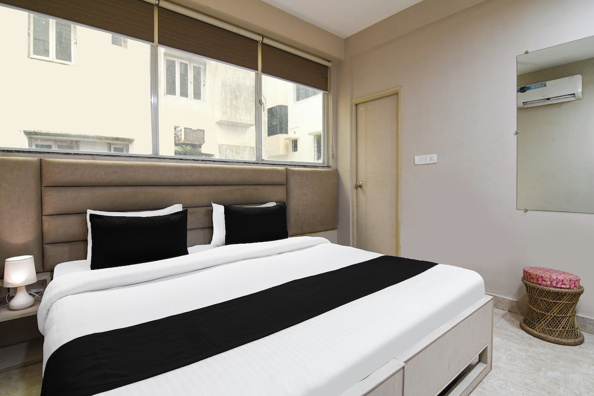 A bedroom with a bed, a nightstand, a lamp, a window with blinds, a door, and a wall-mounted air conditioner.