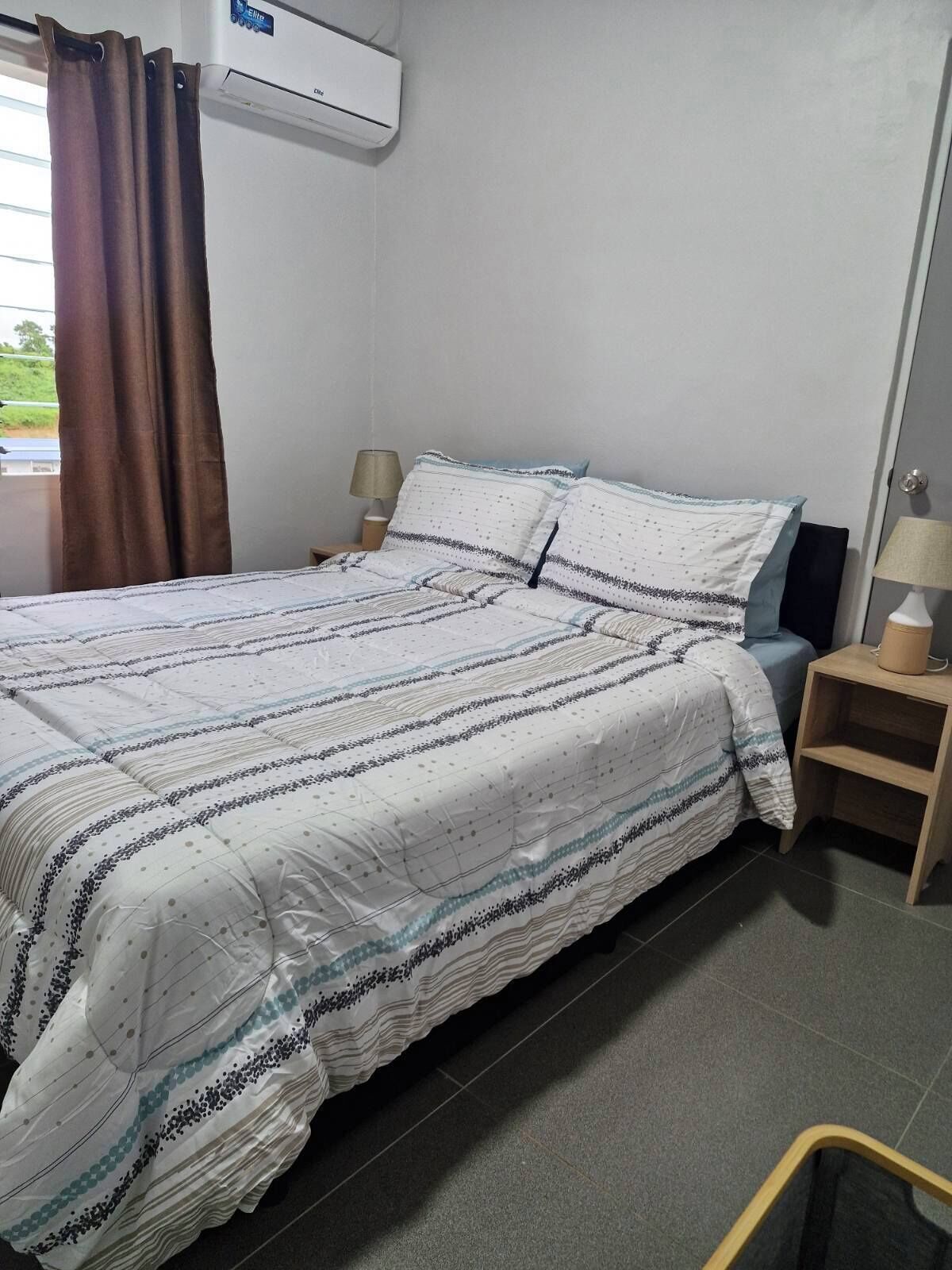 A hotel room with a bed, a nightstand, a lamp, and an air conditioning unit.