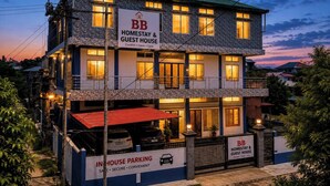 A two-story building with a sign indicating it is a homestay and guest house, featuring a parking area with a red roof.