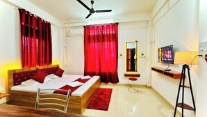 A hotel room with a bed, a desk, a chair, a TV, a ceiling fan, and red curtains. - B.B.Homestay & Guest House (Tezpur)
