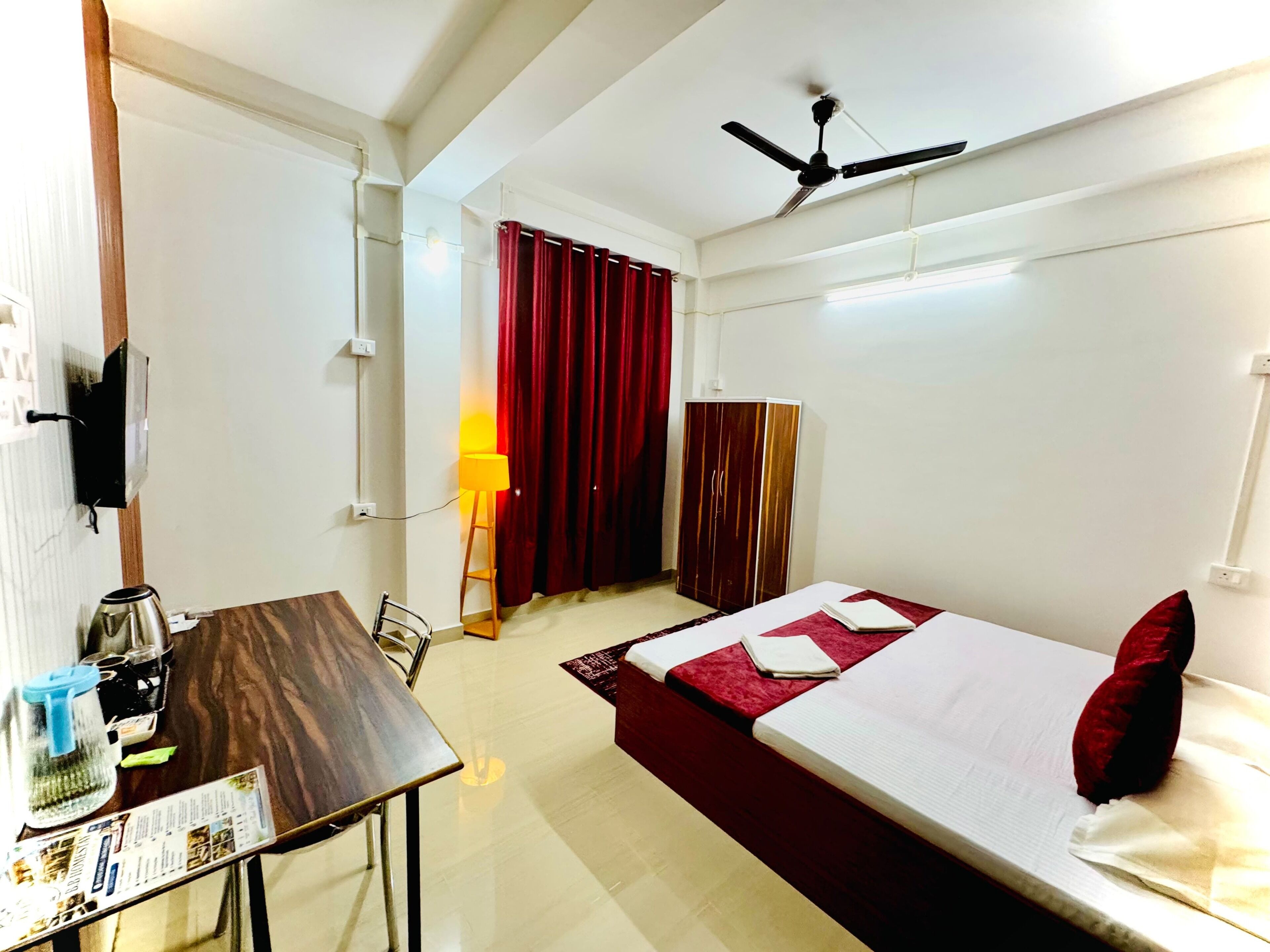 Quarto de hotel com cama, secretária com chaleira, luminária e armário.