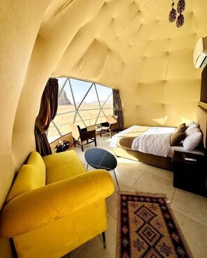 A room with a bed, a yellow armchair, a small table, and a view of the desert through a large window. - Golden STAR luxury Camp (Aqaba)