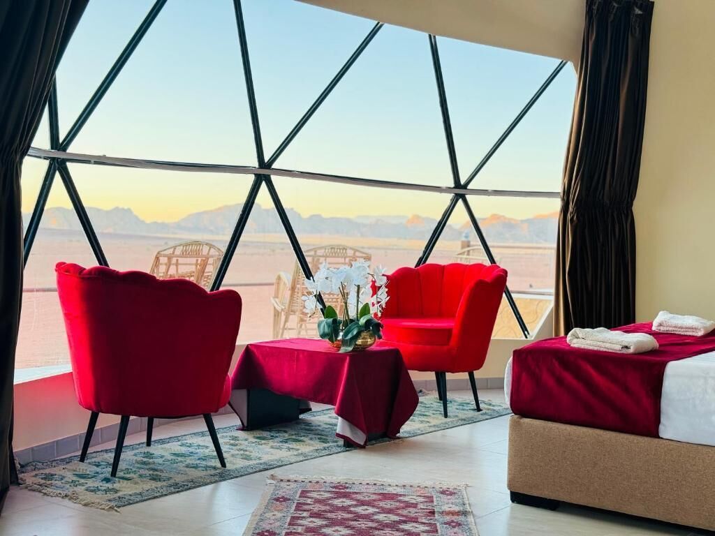 A hotel room with a large window, a bed, two red chairs, a small table with a plant, and a red rug.