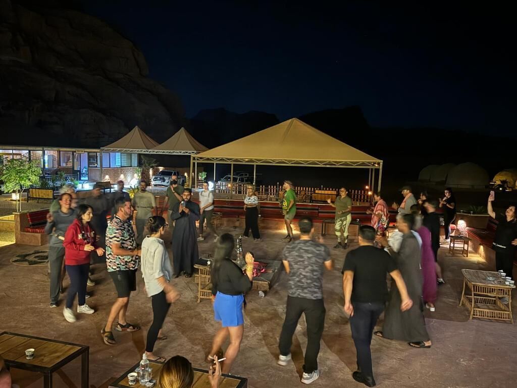 An outdoor gathering area with a tent, tables, and chairs, surrounded by a mountainous landscape.