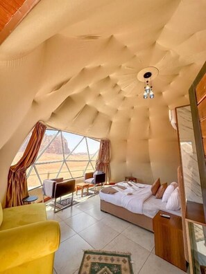 A room with a bed, a yellow armchair, a small table, and a view of the desert through a large window. - Golden STAR luxury Camp (Aqaba)