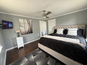A bedroom with a bed, a ceiling fan, a TV mounted on the wall, and a door.