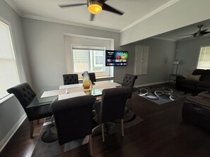 A modern living room with a glass-top dining table, black upholstered chairs, a large flat-screen TV mounted on the wall, and a ceiling fan.