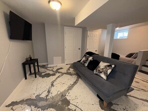 A living room with a grey sofa, a flat-screen TV mounted on the wall, and a white door in the background.