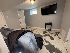 Una habitación con un sofá gris, un televisor de pantalla plana montado en la pared, una mesita con una planta y una puerta que da a otra habitación.