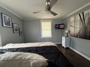 Une chambre à coucher avec un lit, un ventilateur de plafond, une télévision et une fenêtre avec des stores.