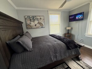 Un dormitorio con cama, un cuadro, televisor y ventana con persianas.