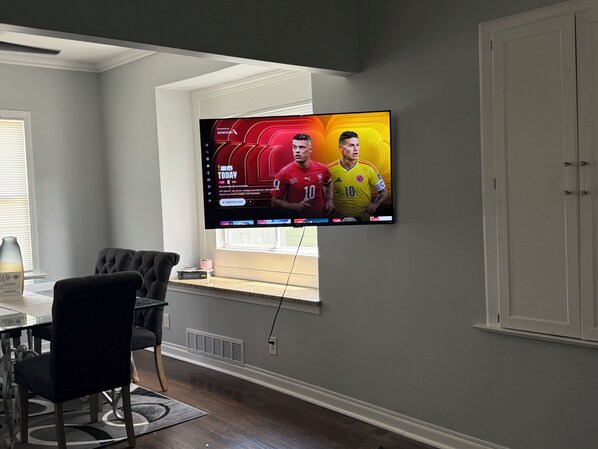 A room with a flat-screen TV mounted on the wall displaying a sports event, a dining table with chairs, and a window with blinds.