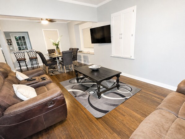 A living room with a brown leather sofa, a dining table with chairs, and a flat-screen TV mounted on the wall.