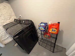 Un espace dédié à la lessive, comprenant un lave-linge, un étagère en métal où sont rangées les bouteilles de lessive, et une boîte de lessive.