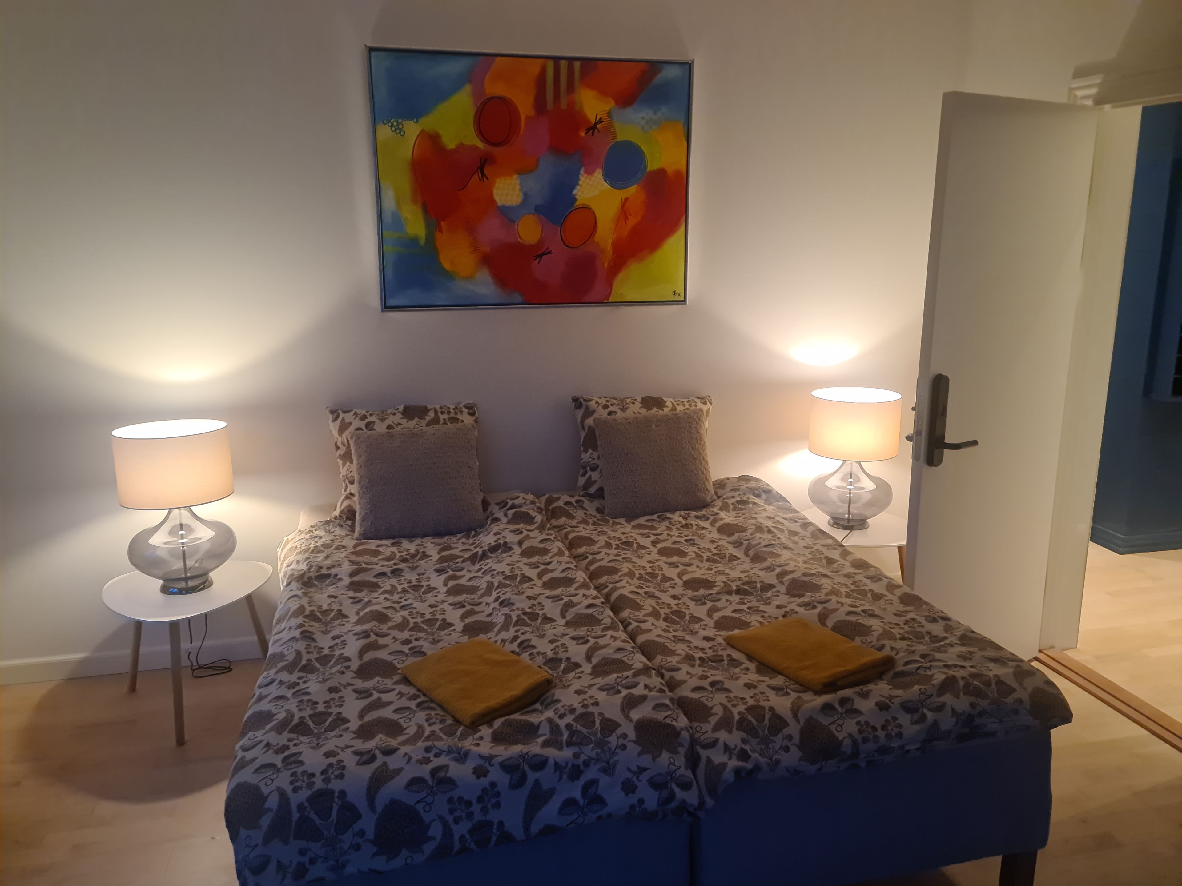 A bedroom with a bed, two bedside lamps, a painting on the wall, and a door.