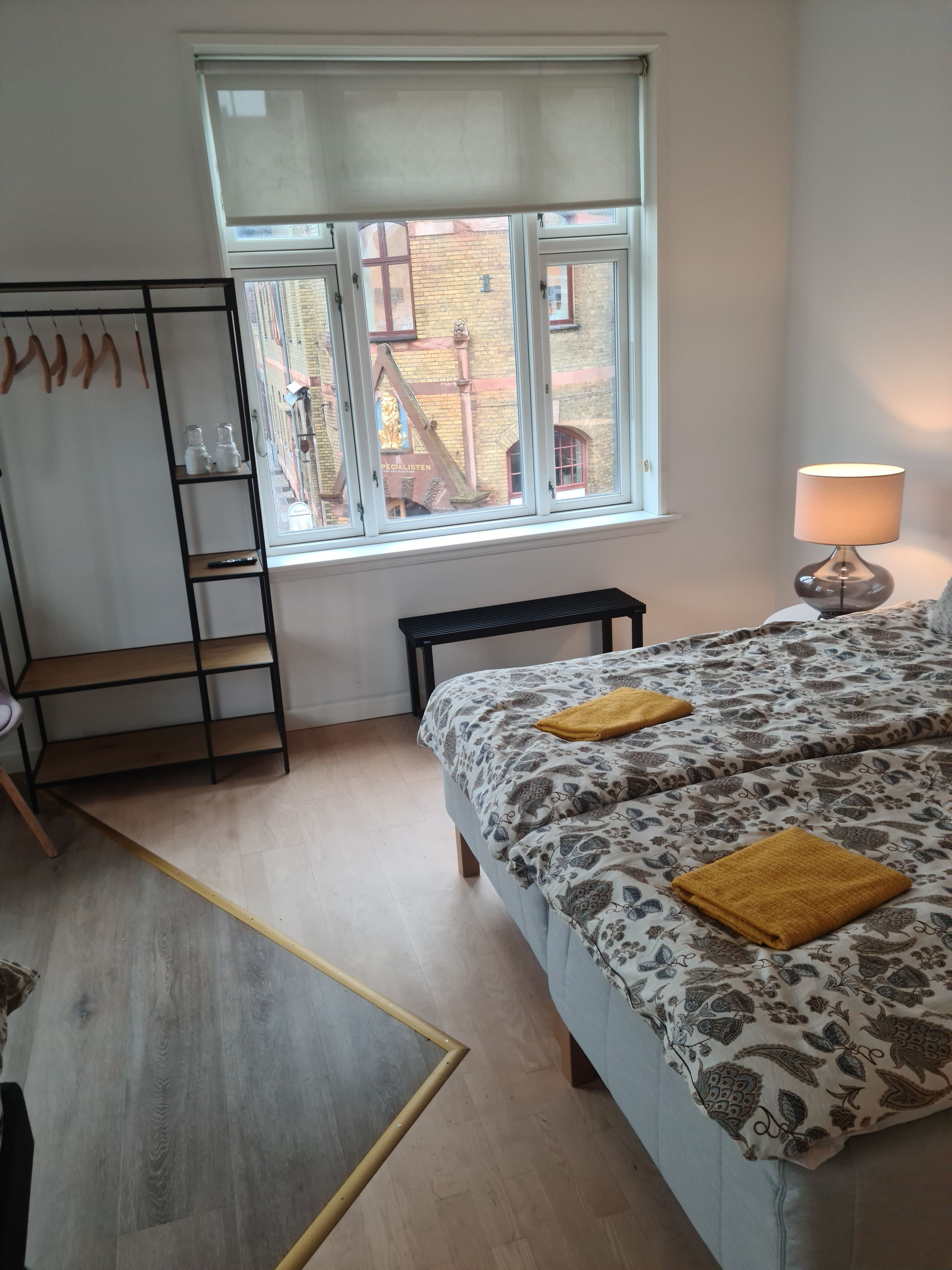 Ein Schlafzimmer mit einem Bett, einem Fenster, einem Nachttisch mit Lampe, Holzboden und einem Kleiderschrank.