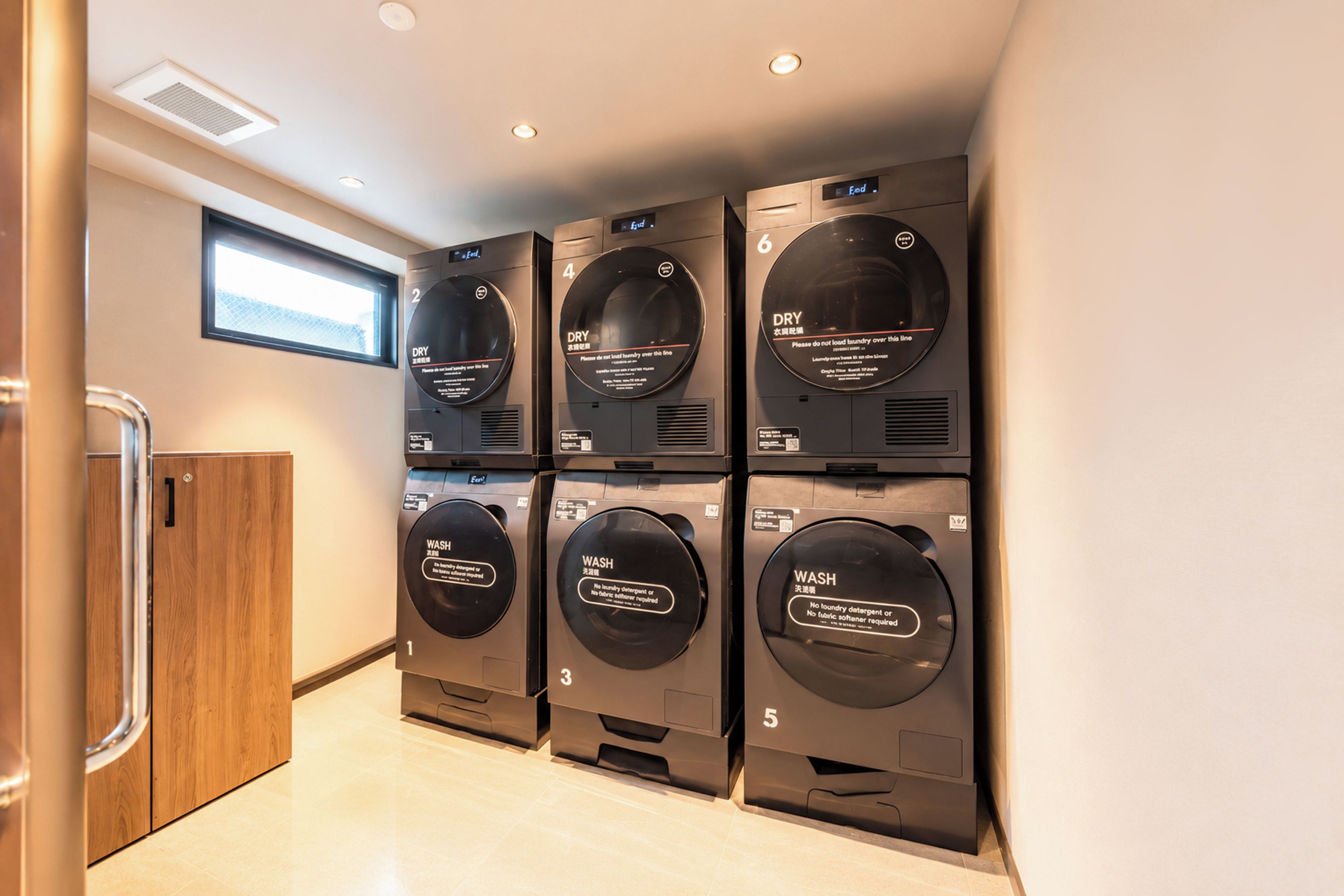 Une rangée de six machines à laver superposées dans une pièce moderne et bien éclairée, avec un meuble en bois sur le côté gauche.