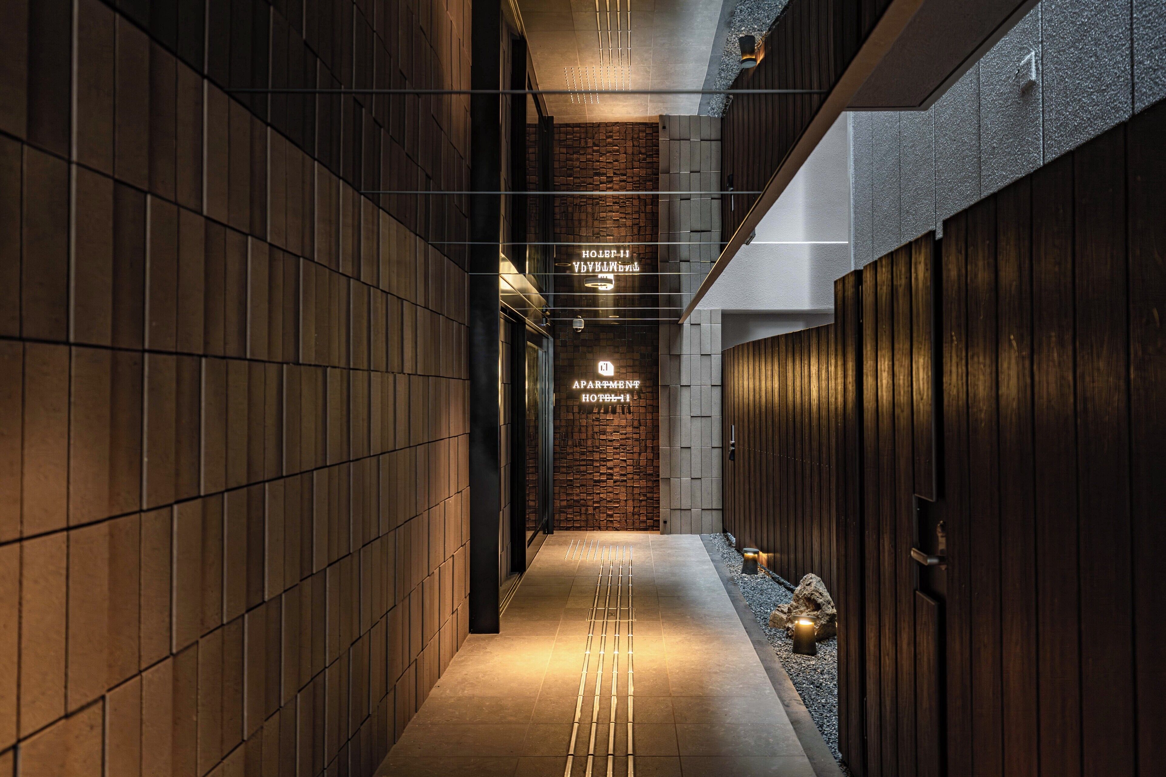 A corridor with a wooden wall on one side and a metal wall on the other, leading to a hotel entrance with a sign.