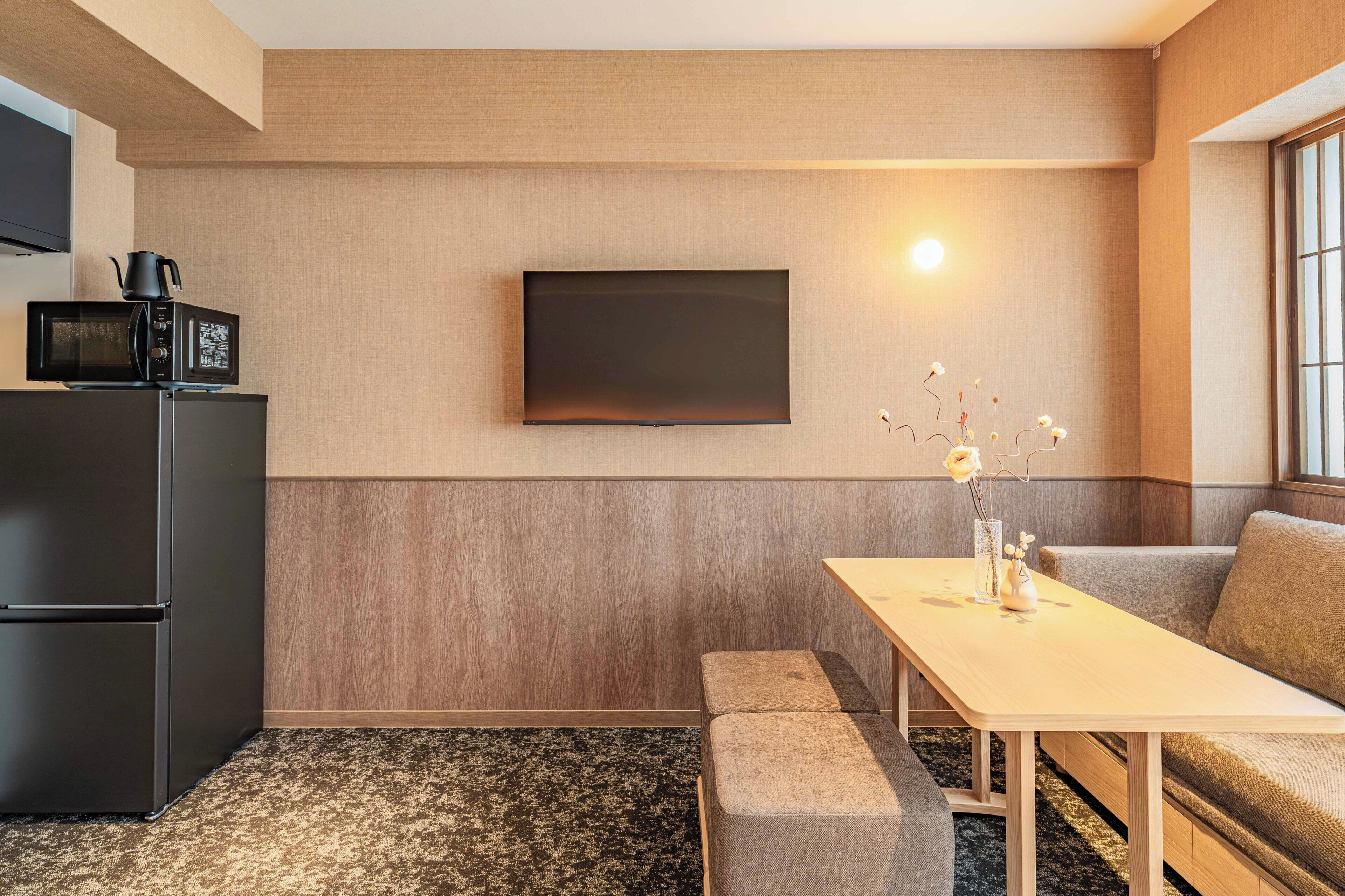 Un espacio habitable compacto con kitchenette, un televisor de pantalla plana montado en la pared, una pequeña mesa de comedor con un florero y un sofá.