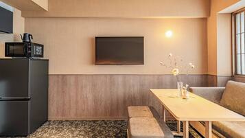 A compact living space with a kitchenette, a flat-screen TV mounted on the wall, a small dining table with a vase of flowers, and a sofa.