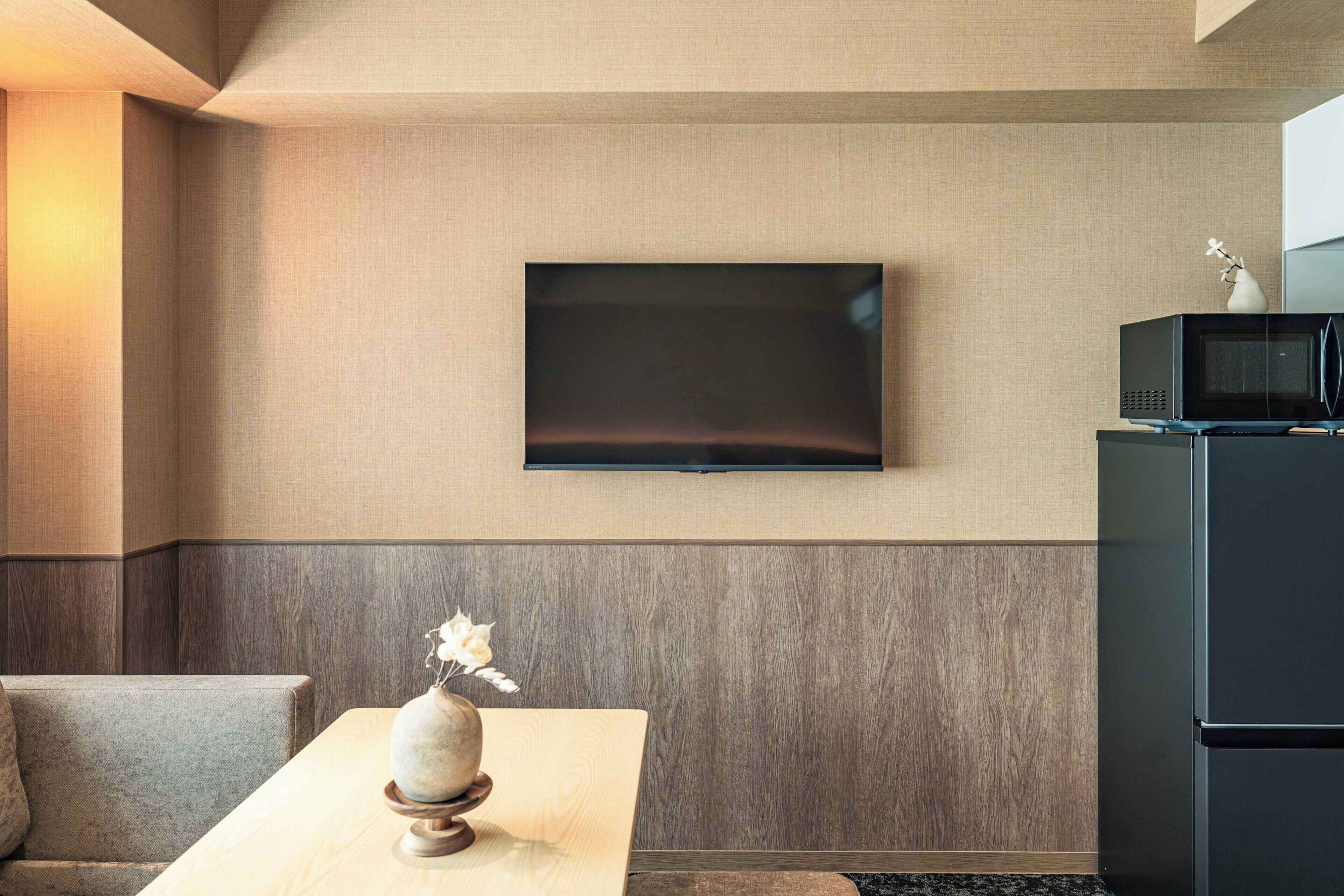 Una sala de estar moderna con un televisor de pantalla plana montado en la pared, un sofá de color oscuro, una mesa de centro de madera con un jarrón decorativo y un microondas negro sobre un mueble.