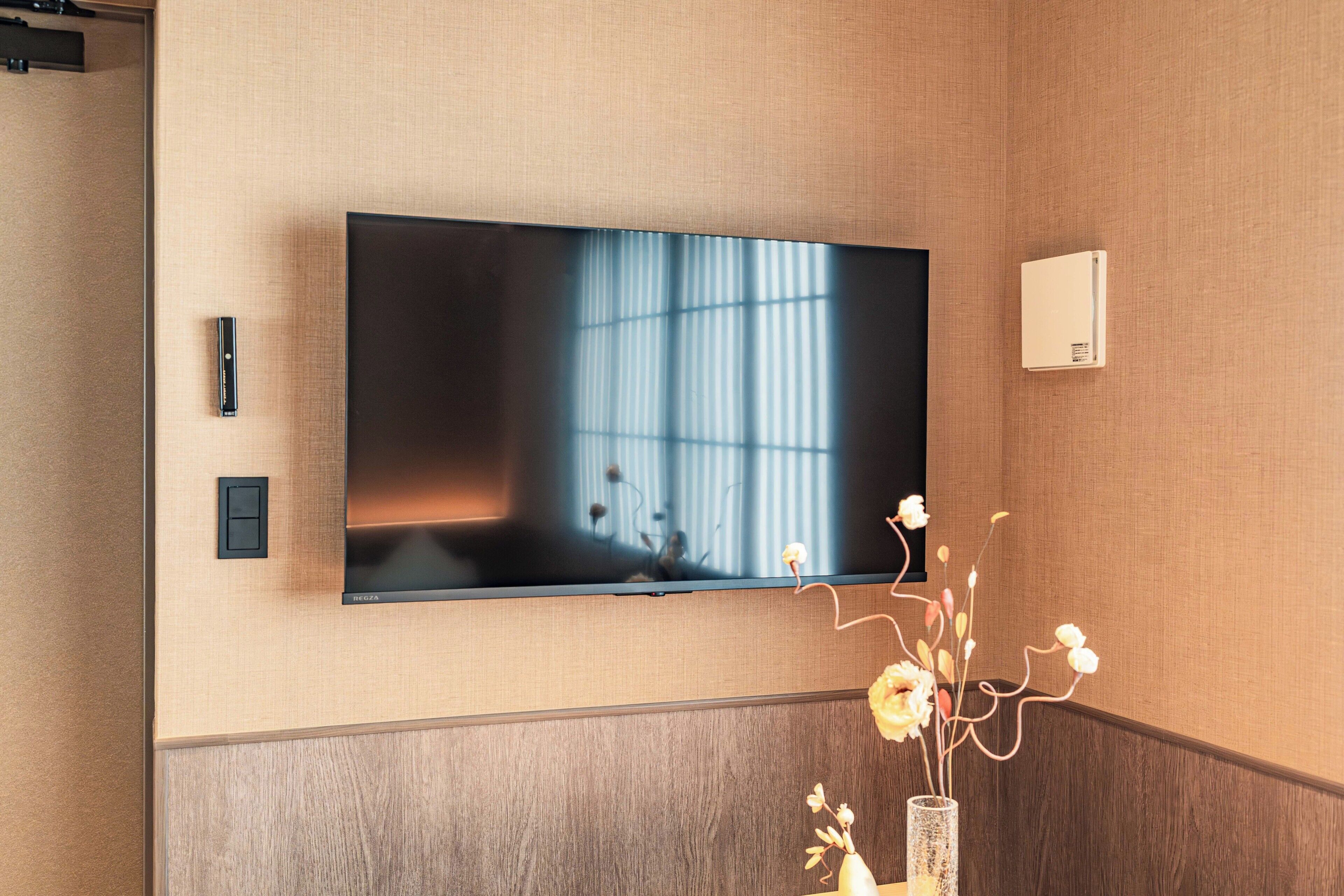 A flat-screen TV mounted on a wall with a wooden panel design, a remote control, and a thermostat on the wall.