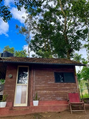 A wooden house with a tiled roof, a door, and a window. There are potted plants and a chair outside. - Happy Feet Wayanad (Sulthan Bathery)