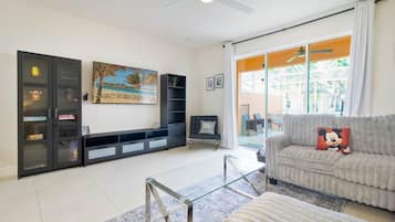 A living room with a sofa, a glass coffee table, a bookshelf, a painting, and a sliding glass door leading to an outdoor area.
