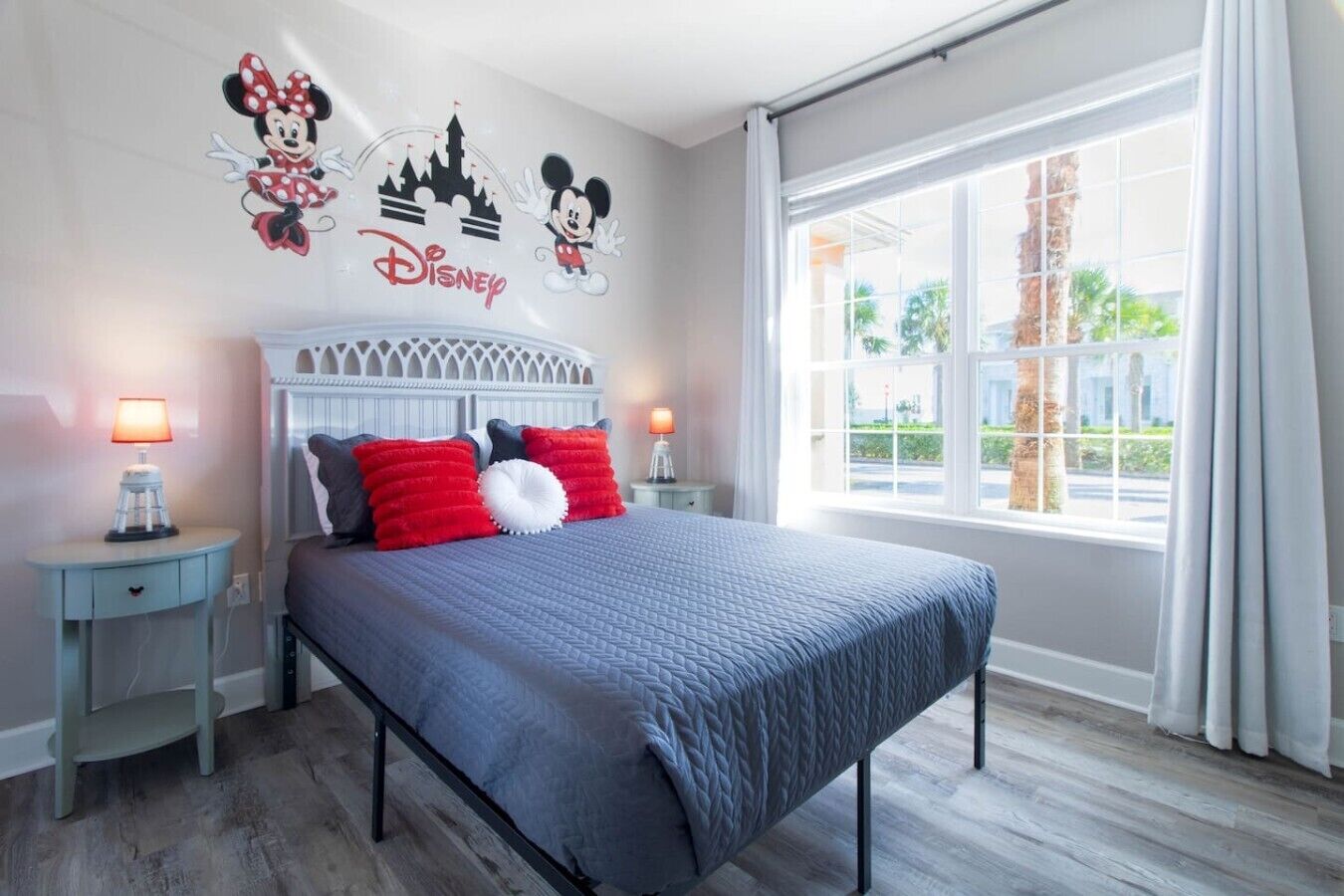 Ein Hotelzimmer mit einem Bett, zwei Nachttischen mit Lampen, einem Fenster mit Vorhängen und Wandbildern mit Disney-Motiven.