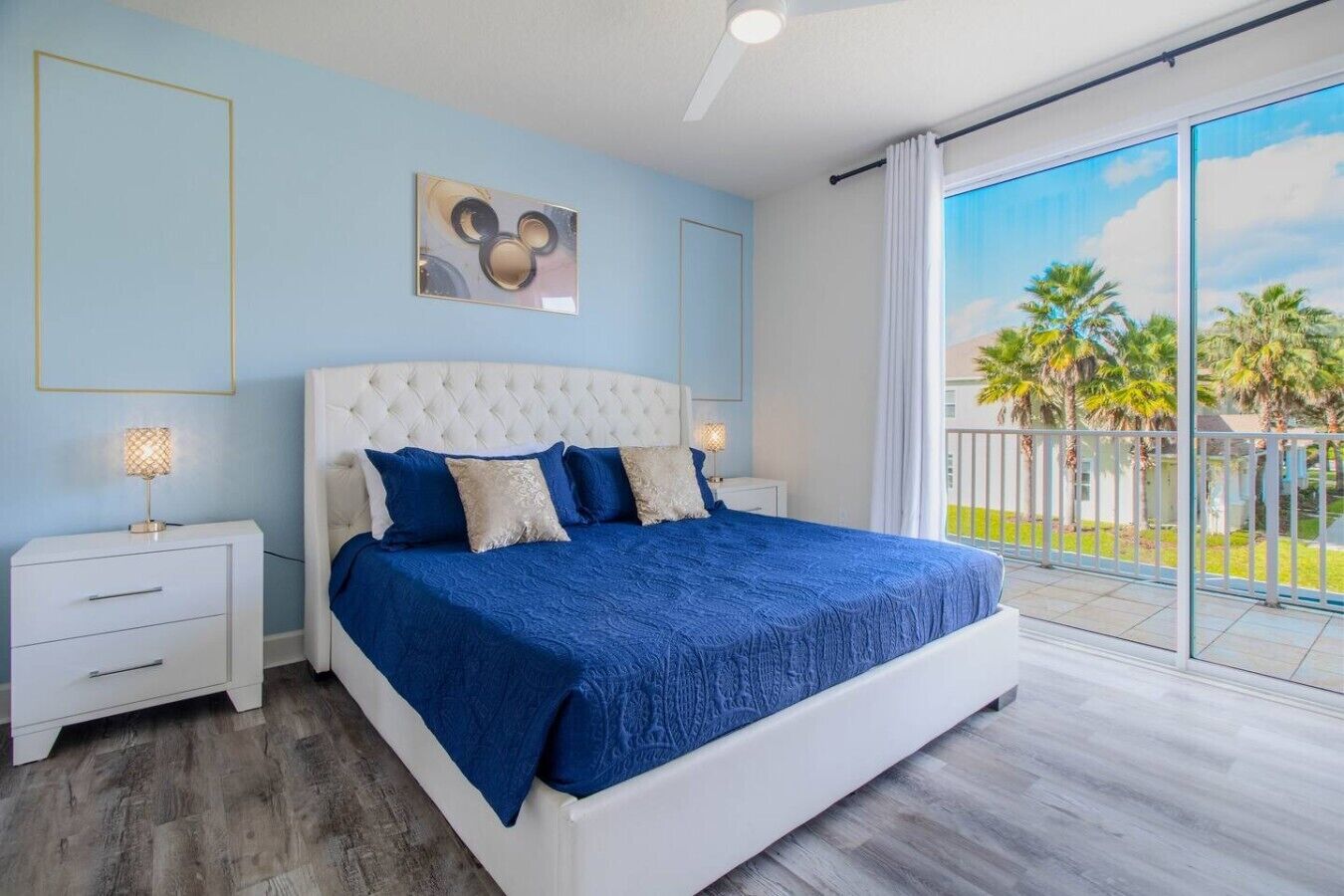 A bedroom with a large bed, a white headboard, and a view of palm trees through large windows.