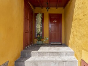 Entrada de um edifício com parede amarela, duas portas de madeira, uma placa de boas-vindas, um vaso de planta e uma bicicleta.