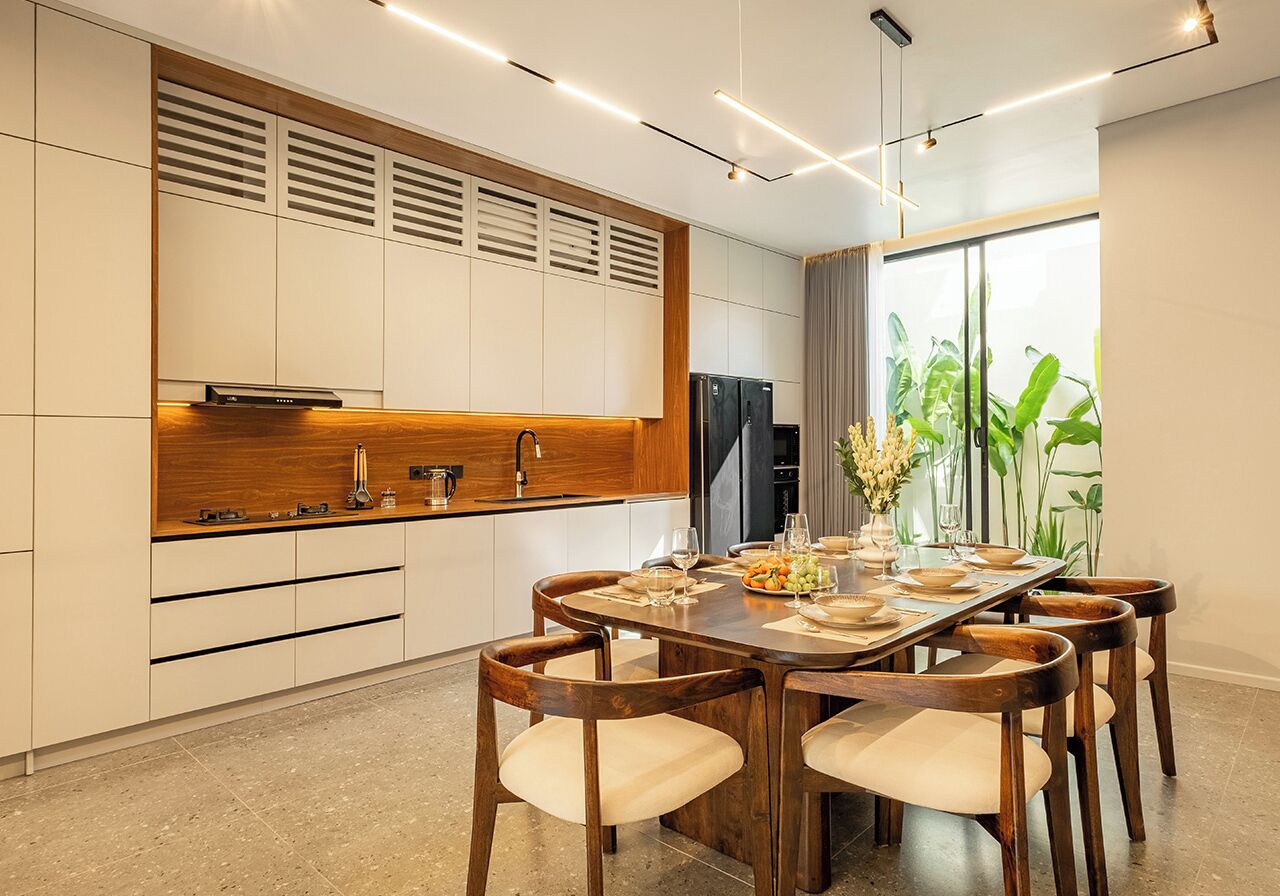 A modern kitchen with a dining area, featuring a wooden table and chairs, a built-in oven, and a large window with plants.