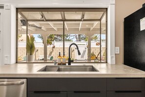 A kitchen with a modern design, featuring a stainless steel sink, a black faucet, and a window that offers a view of the outdoors. - 1 Step to the Park - 6BR • Game On! Arcade • BBQ • Pool Table - Long Stay Luxury (Huntington Beach)